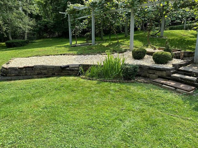 A raised garden bed filled with light-colored gravel, featuring a stone retaining wall and stone steps in a grassy yard.