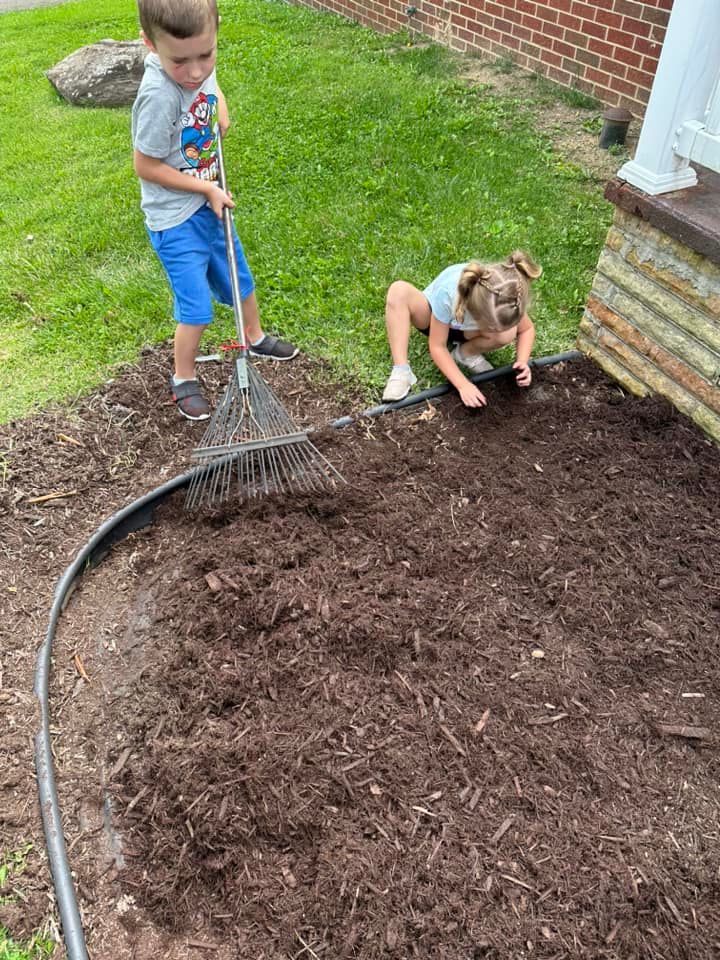Two children spread dark mulch in a yard garden bed; one rakes while the other uses their hands.