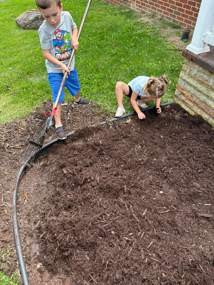 Two children work in a garden bed with fresh brown mulch, one using a metal rake and the other kneeling to spread it.
