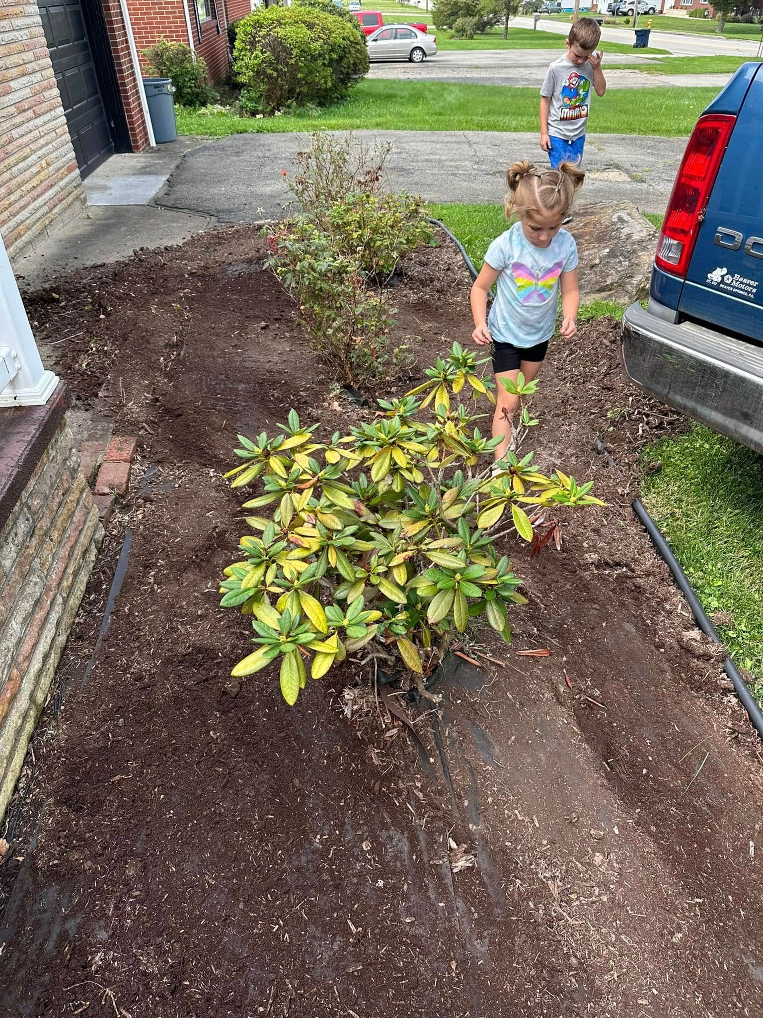 Two children explore a mulched garden bed in front of a brick house, next to a parked blue vehicle.