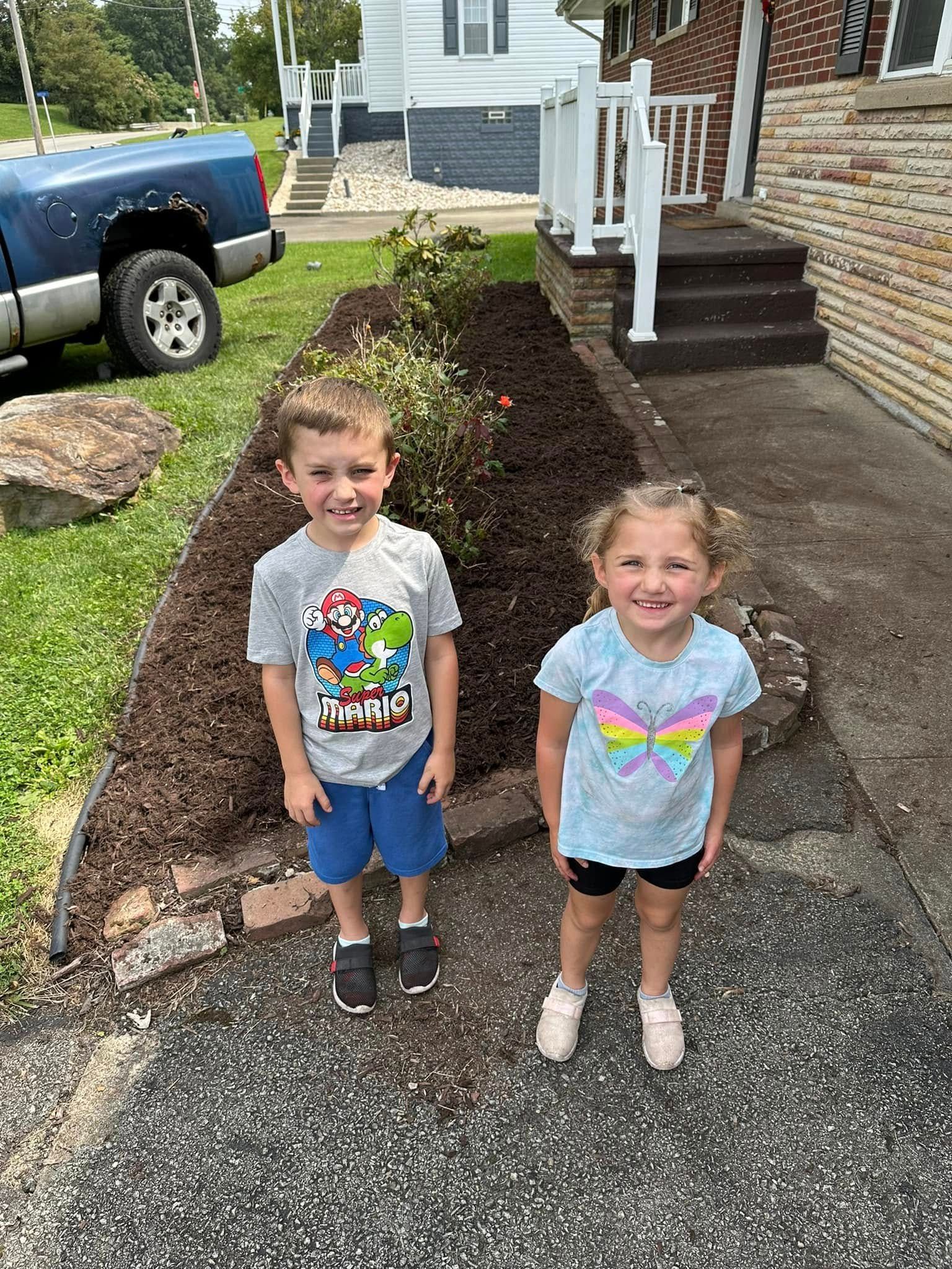 Two children standing outside on a sunny day next to a mulched garden bed and a blue truck.