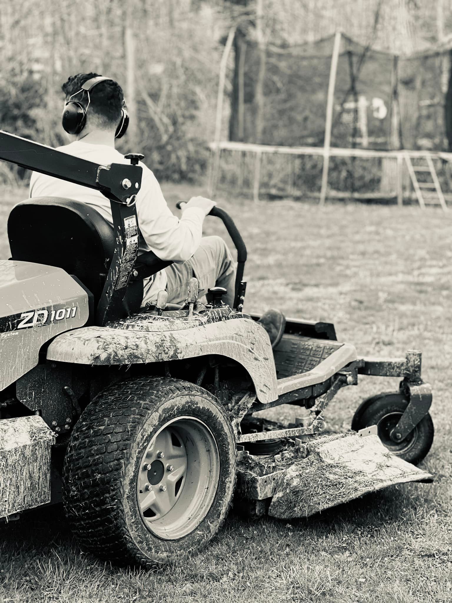 A person wearing hearing protection operates a muddy zero-turn mower in a yard near a trampoline.