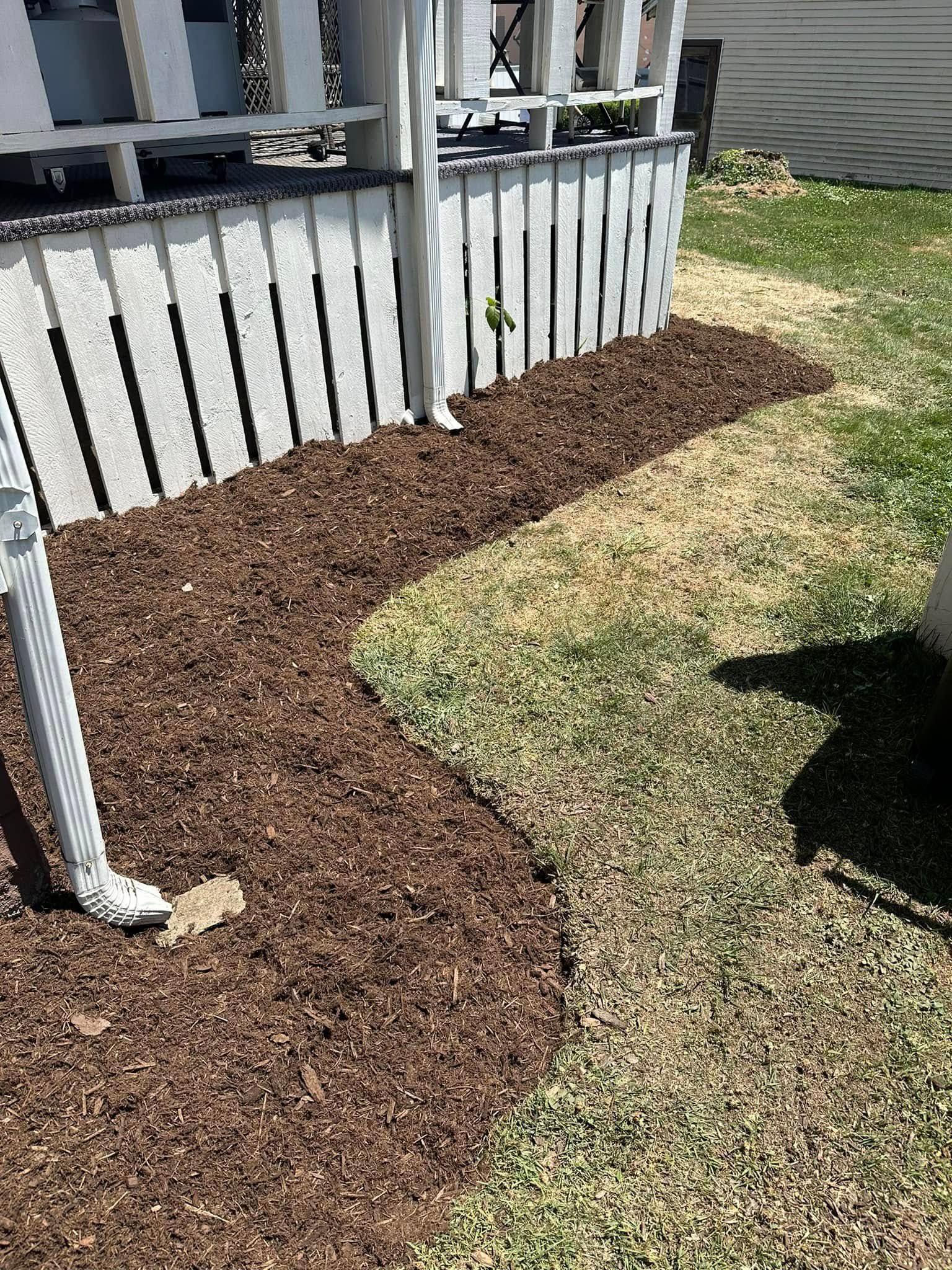 A curved garden bed filled with dark brown wood mulch sits in front of a white latticed deck with a white downspout.