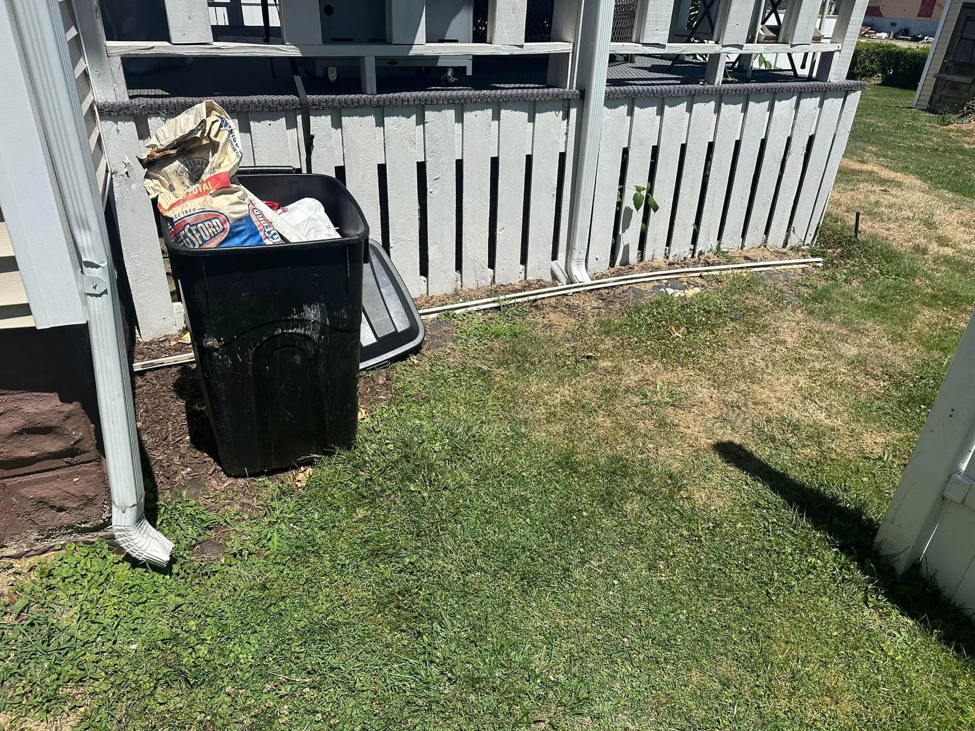 A black trash can filled with bags sits outside on a lawn, next to a white wooden porch fence and a drain pipe.
