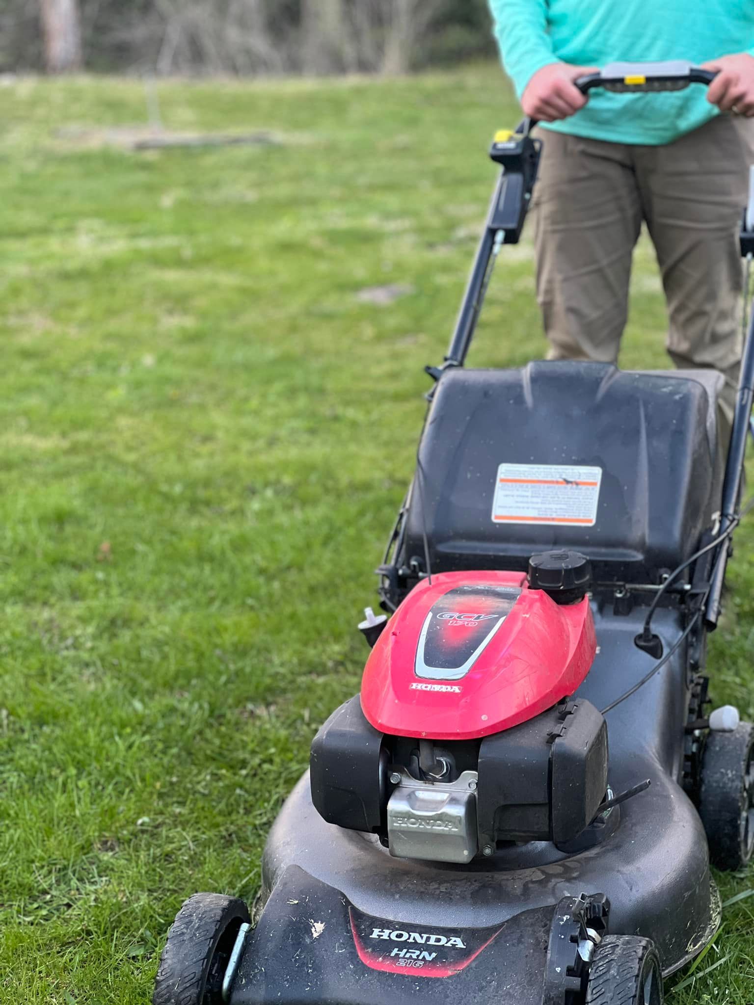 A person in a light blue long-sleeved shirt and tan pants pushes a black and red Honda lawnmower across a grassy yard.