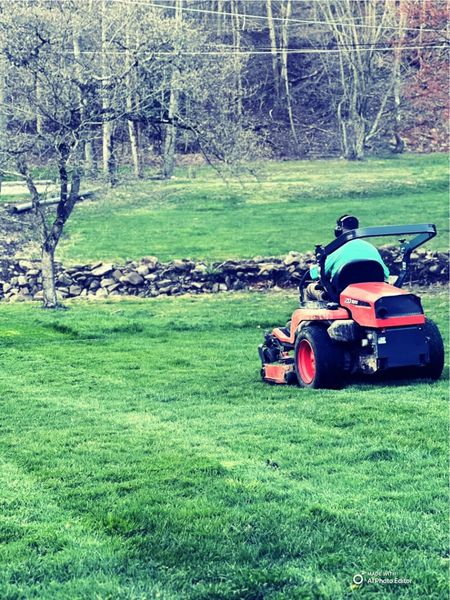 A person wearing a bright turquoise shirt rides an orange zero-turn mower across a green lawn near a stone wall and tree.