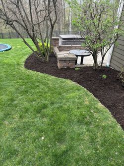 A stone patio area with a table and bench is surrounded by dark mulch and trees in a grassy backyard.