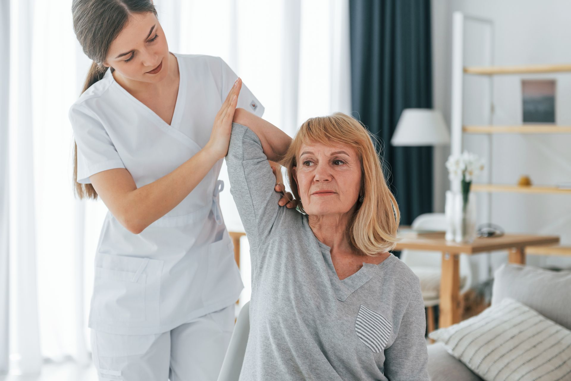 Woman in white uniform assisting elderly woman with arm exercise indoors.