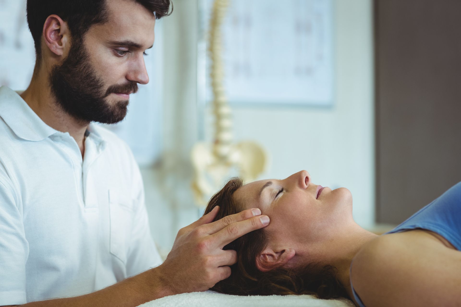 Man in white shirt gently touching woman's forehead. Setting: medical office, spine model visible.