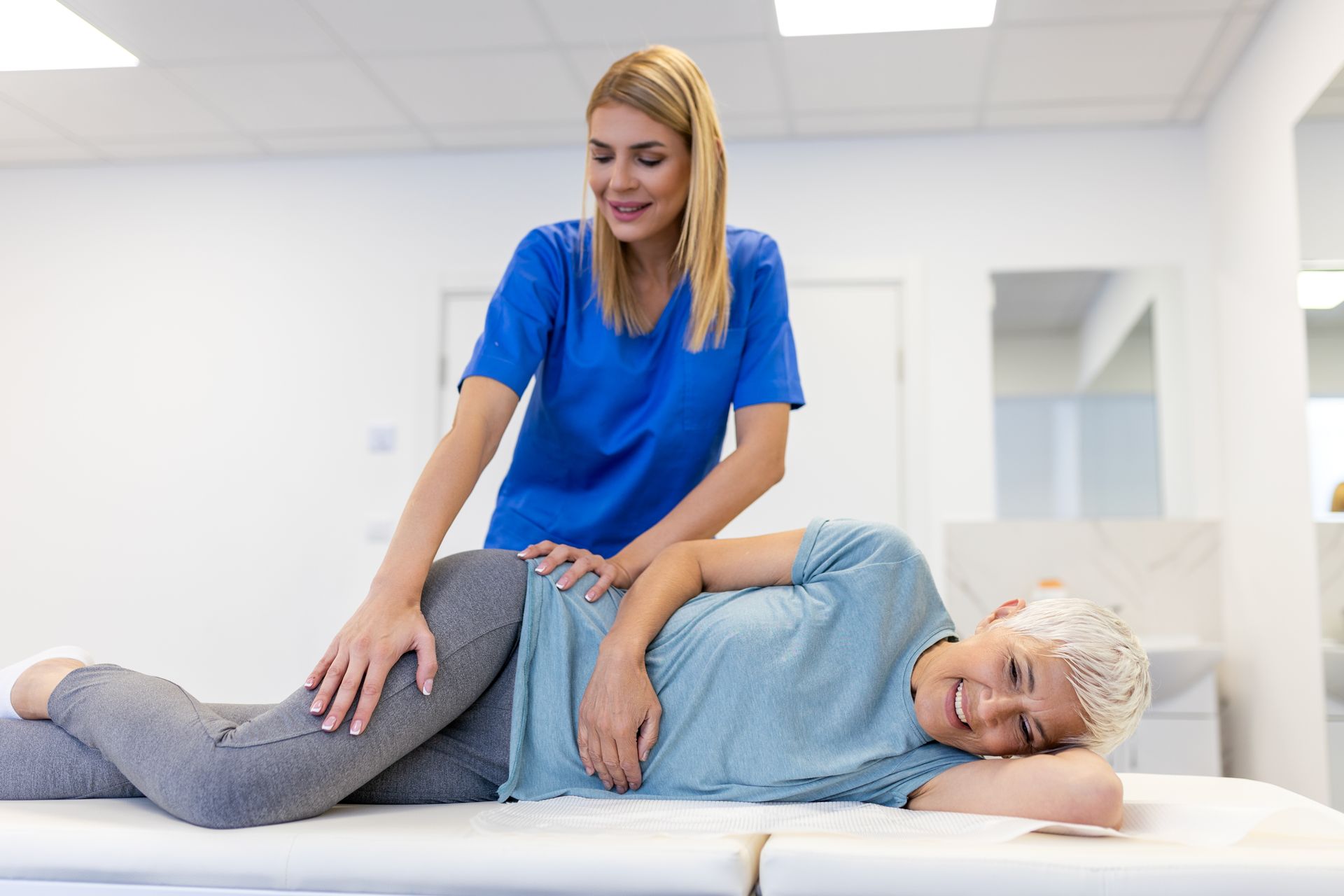 Physiotherapist in blue scrubs, assisting a smiling woman in gray on a treatment table.