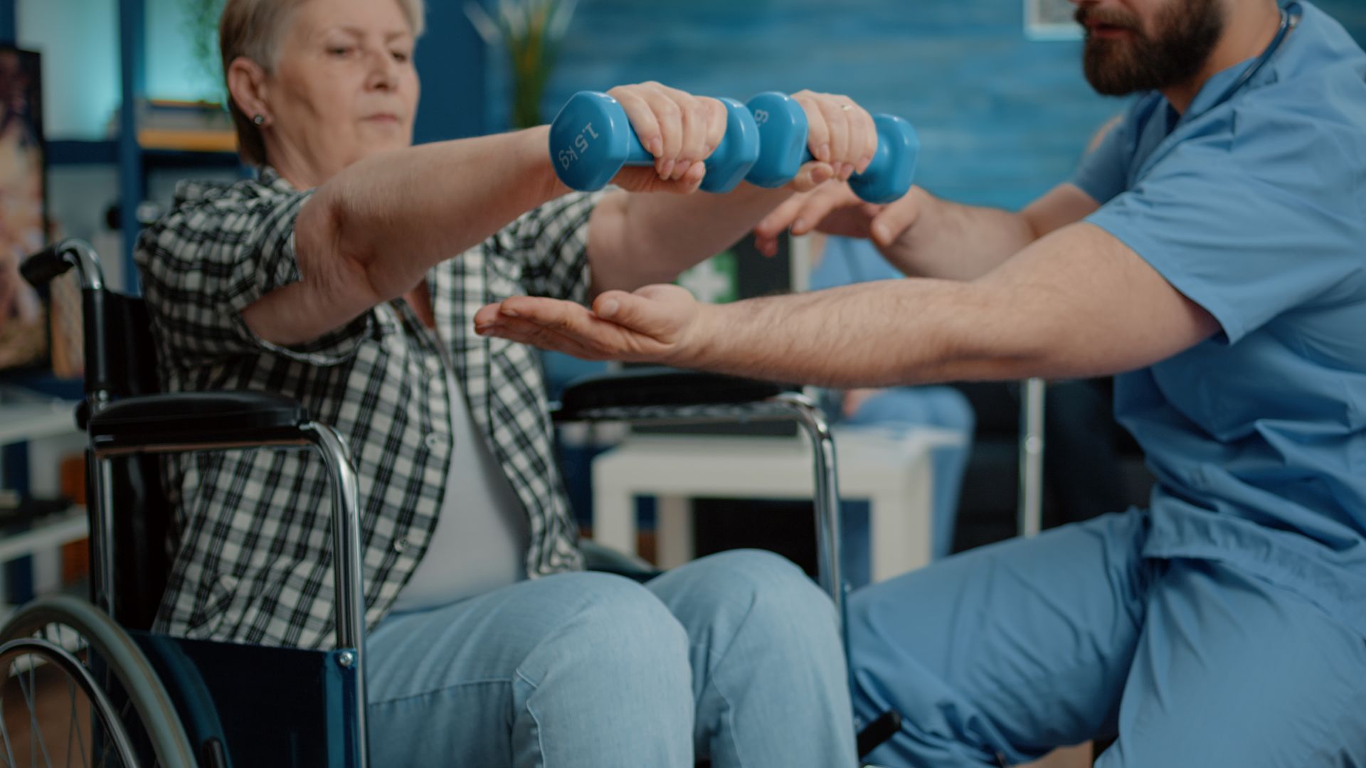 An elderly woman in a wheelchair exercises with small weights, guided by a male healthcare worker.