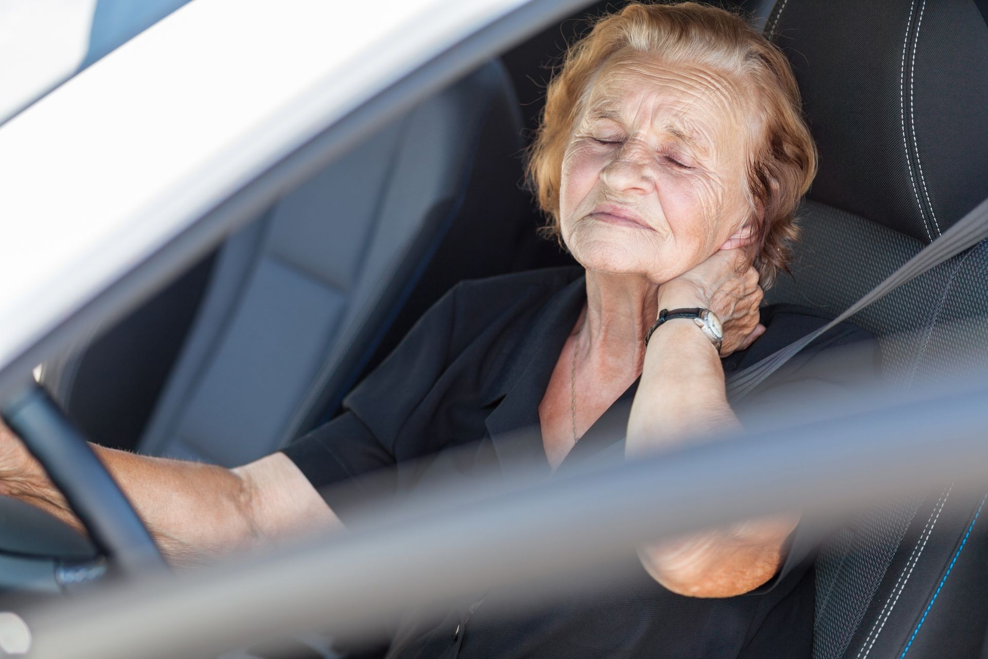 Elderly woman in car, holding neck, looking distressed.