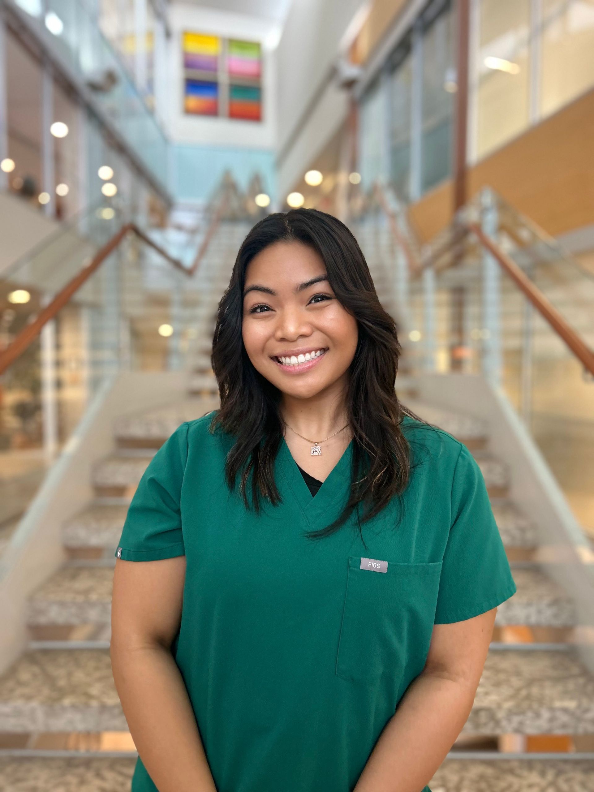 Woman in green scrubs smiling, standing in front of a staircase.