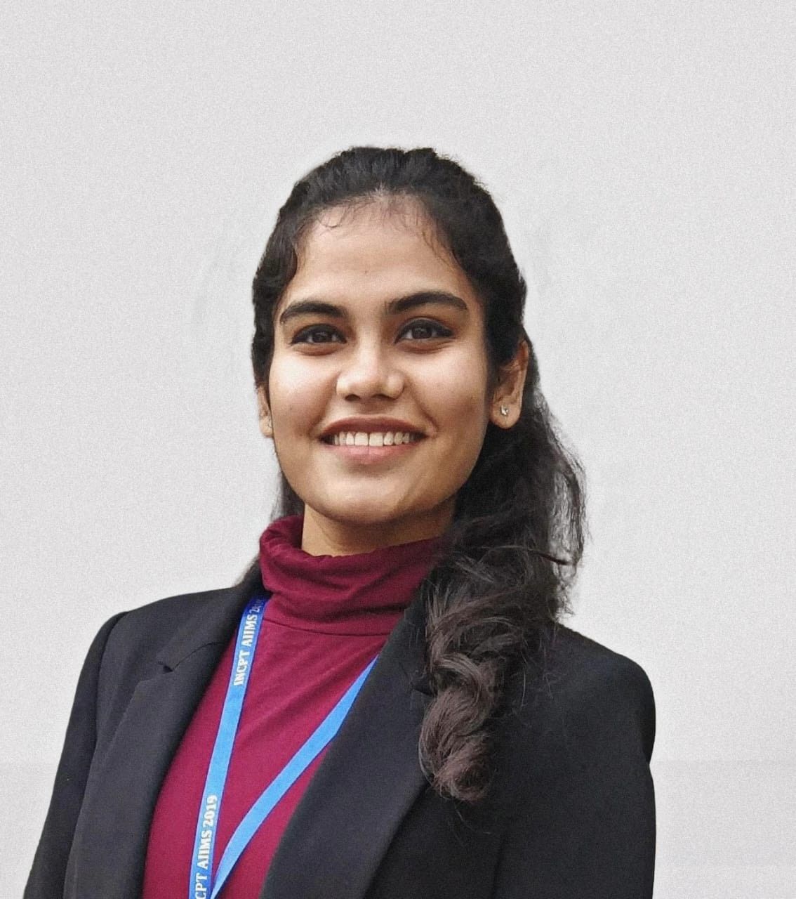 Young woman with dark hair smiling, wearing a maroon turtleneck, black blazer, and a lanyard against a light background.
