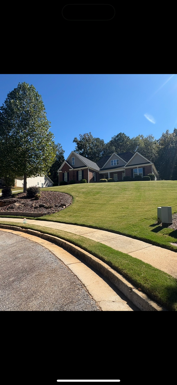 A concrete irrigation canal runs through green rice fields, with houses in the background under a cloudy sky.