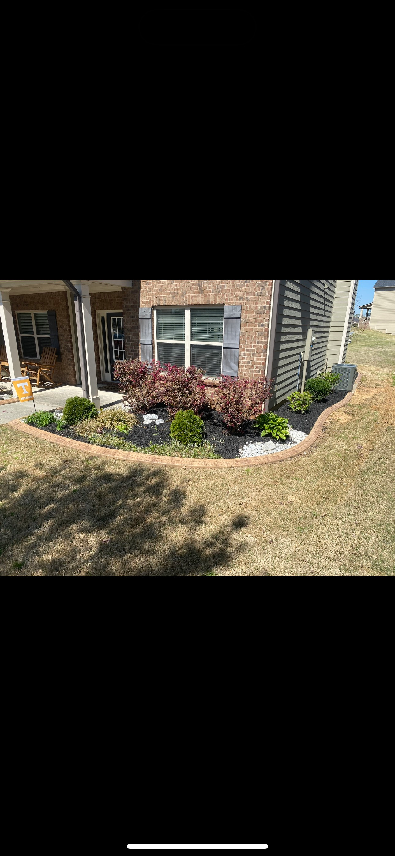 A brick house with landscaping, bushes and greenery along the front, and a curved lawn.
