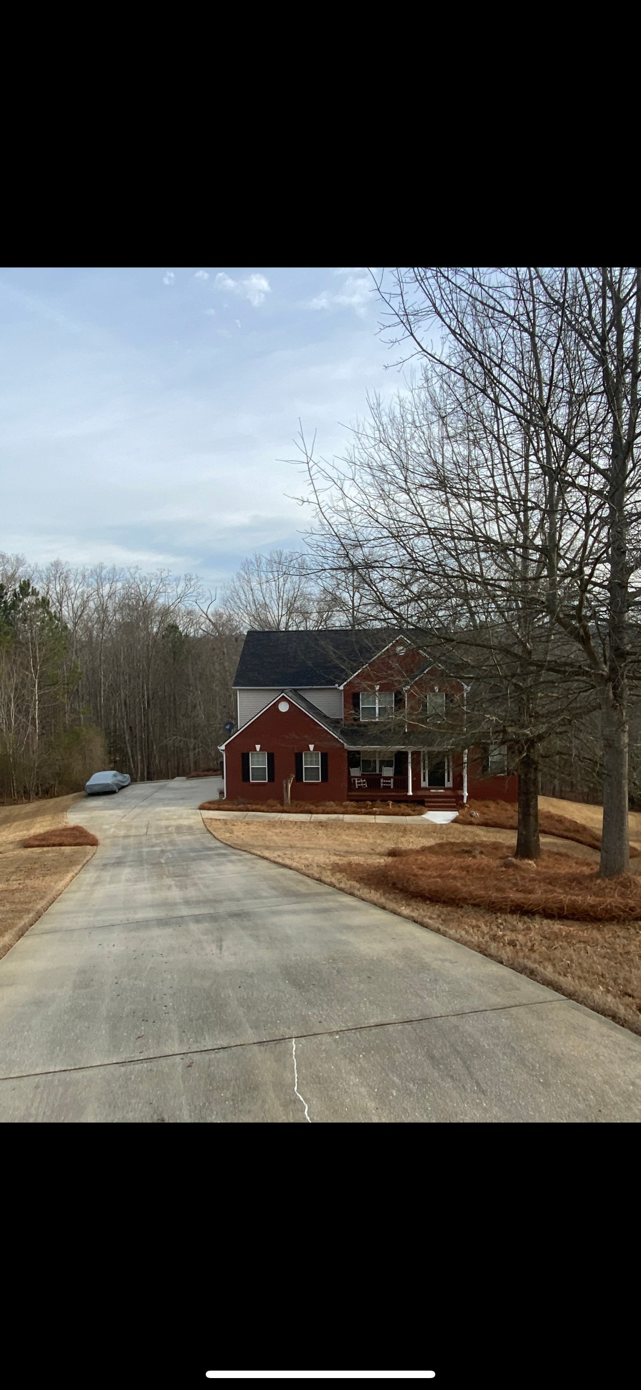 Long driveway leading to a two-story brick house surrounded by trees. Cloudy sky.