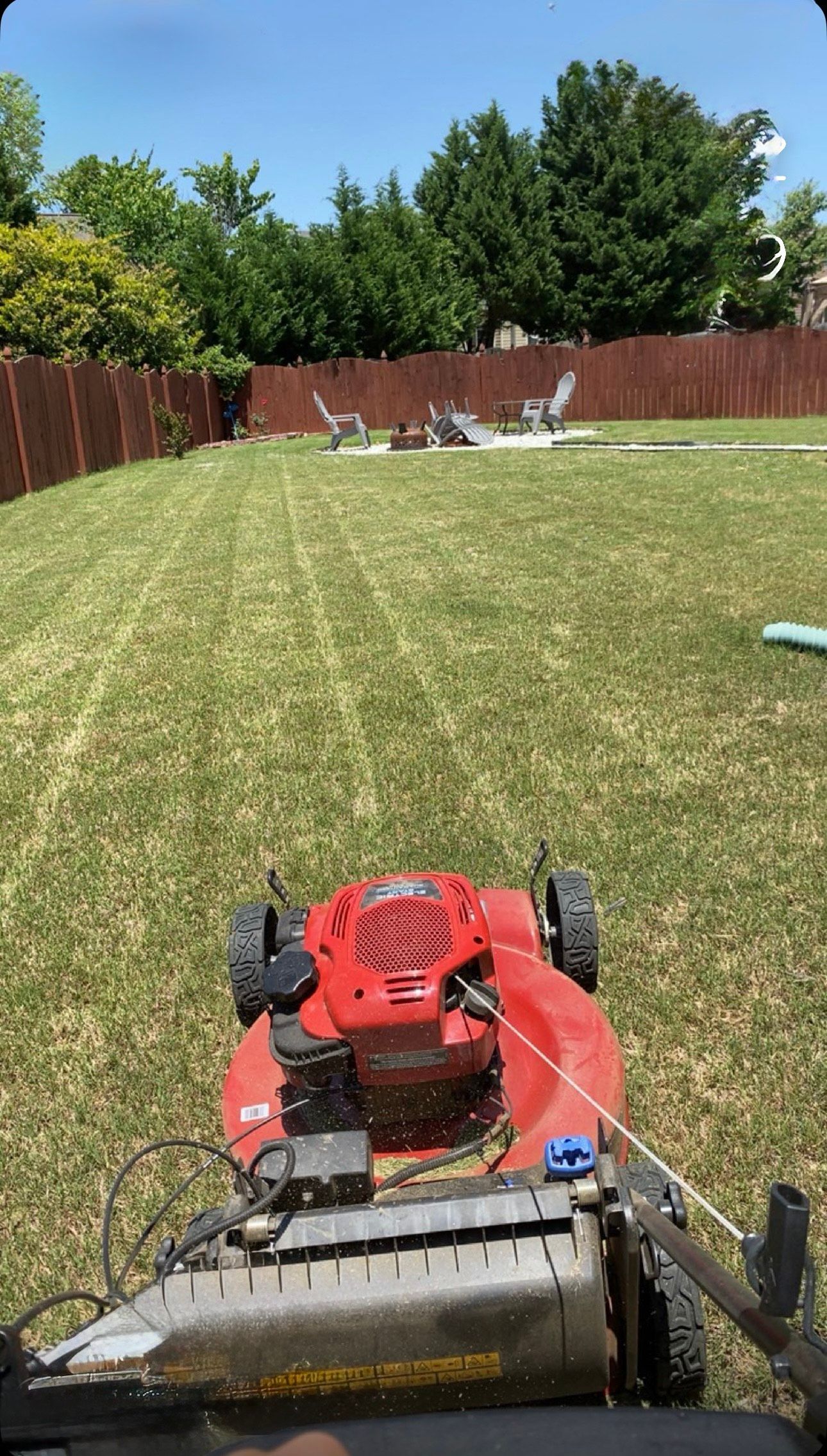 Red lawnmower cutting grass in a backyard with a wooden fence and trees.