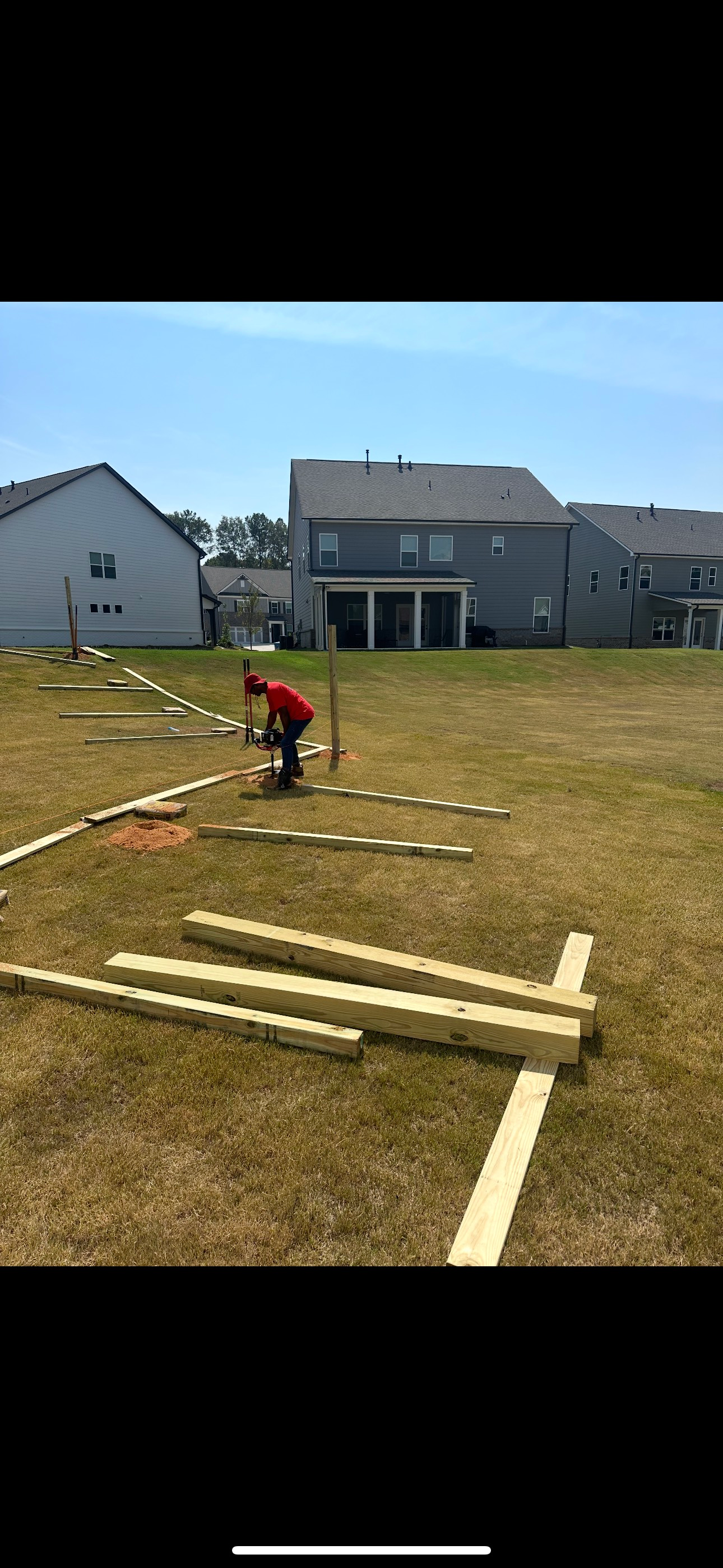 Person working on construction in a grassy backyard with lumber, stakes, and houses in the background. Sunny day.
