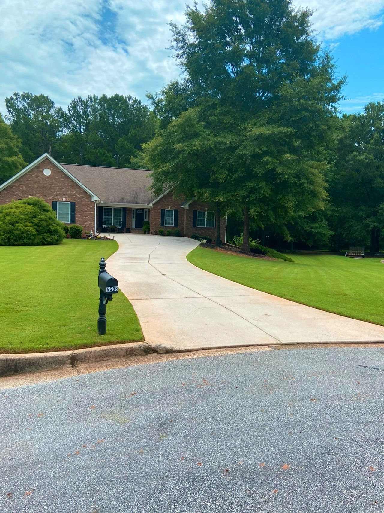 Brick house with long driveway, mailbox, and lush green lawn under a partly cloudy sky.