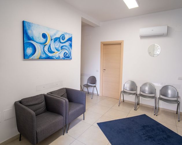 Una sala d'attesa con due poltrone grigie, tre sedie di metallo, un tappeto blu e un quadro astratto blu su una parete bianca.