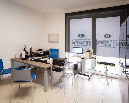 Uno studio medico con una scrivania, due sedie blu e attrezzature oftalmiche davanti a una finestra con tende di marca.