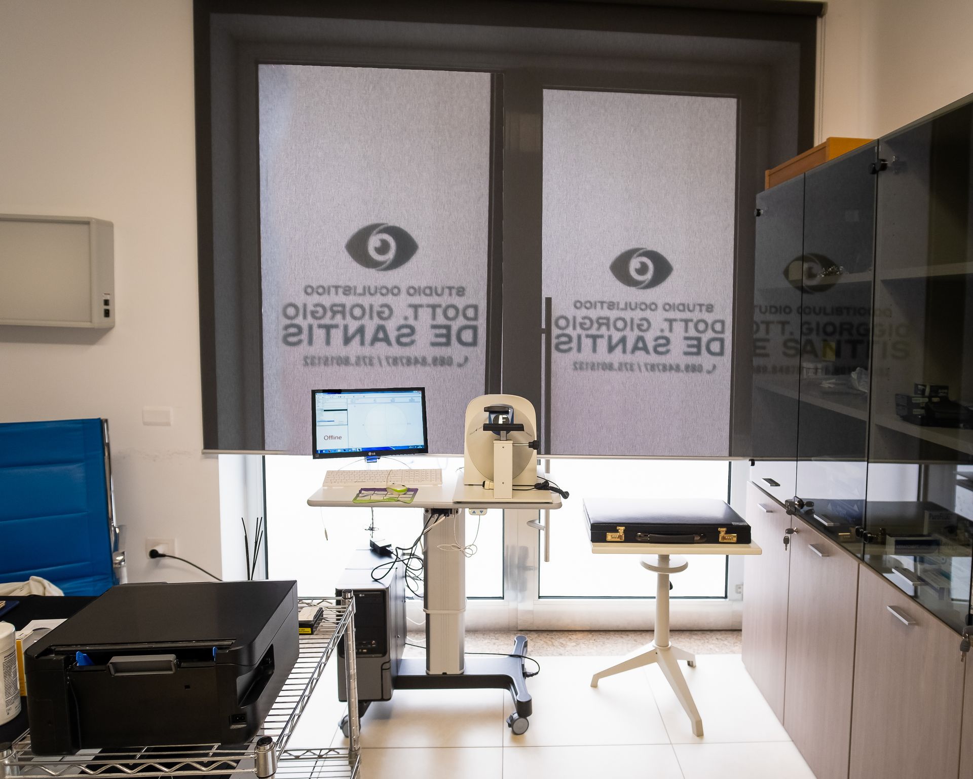 Uno studio oculistico con una scrivania per l'apparecchio per l'esame della vista, un computer, una sedia e armadietti medici.