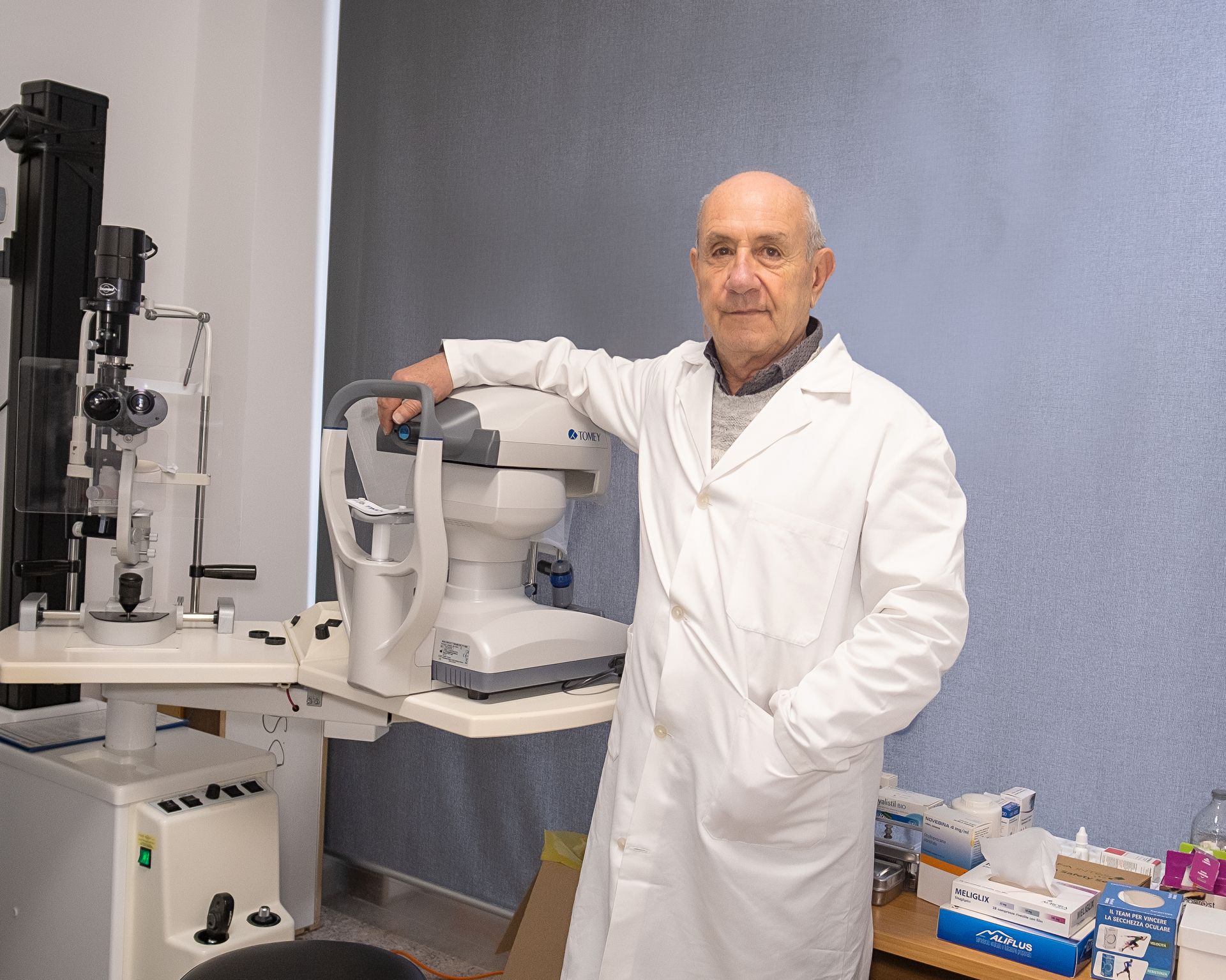 Un professionista sanitario in camice bianco posa accanto a un dispositivo diagnostico oftalmico in un ambulatorio.