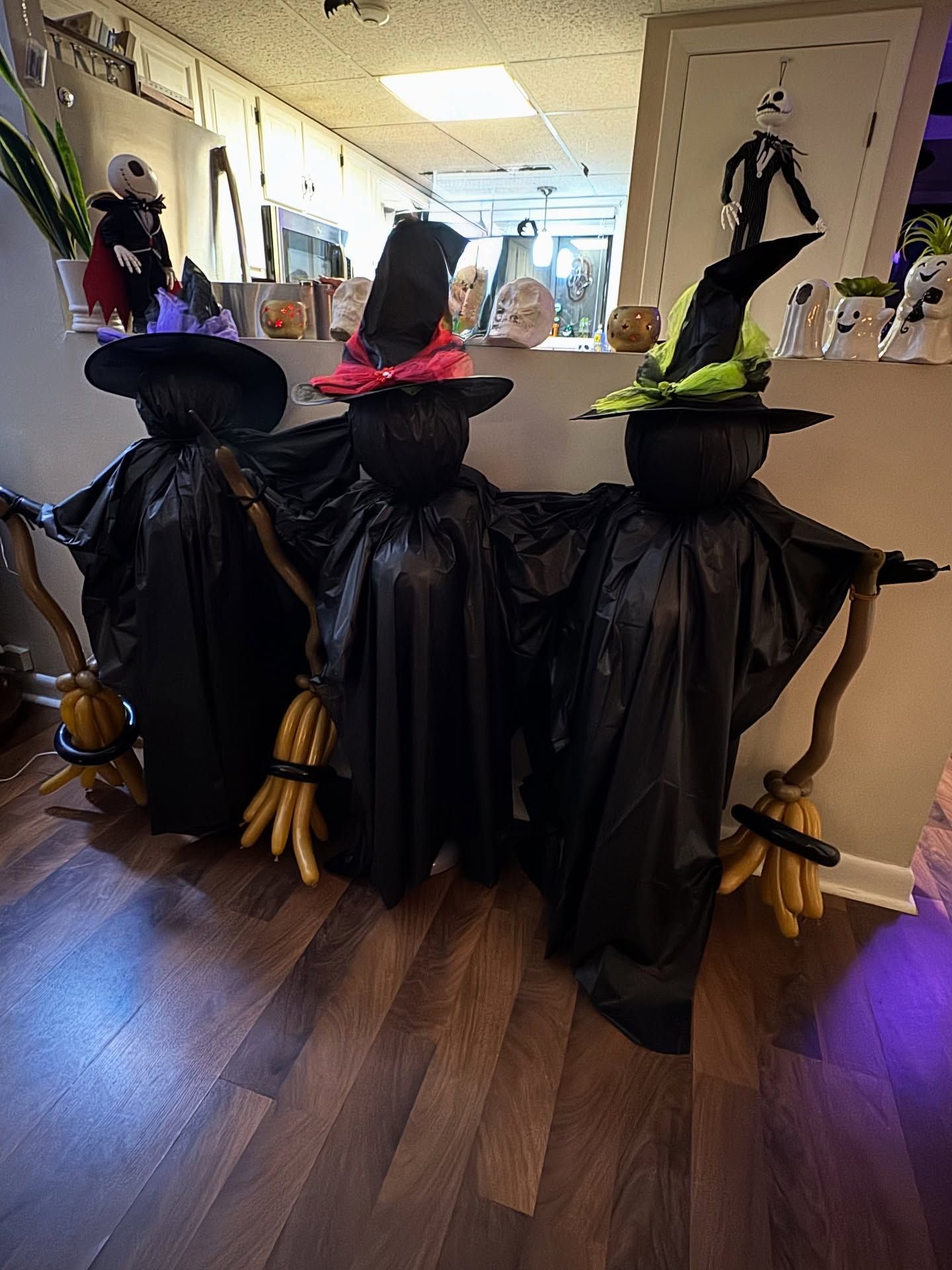 Three Halloween witch mannequins in black cloaks stand in a hallway, with colorful hats and brooms.