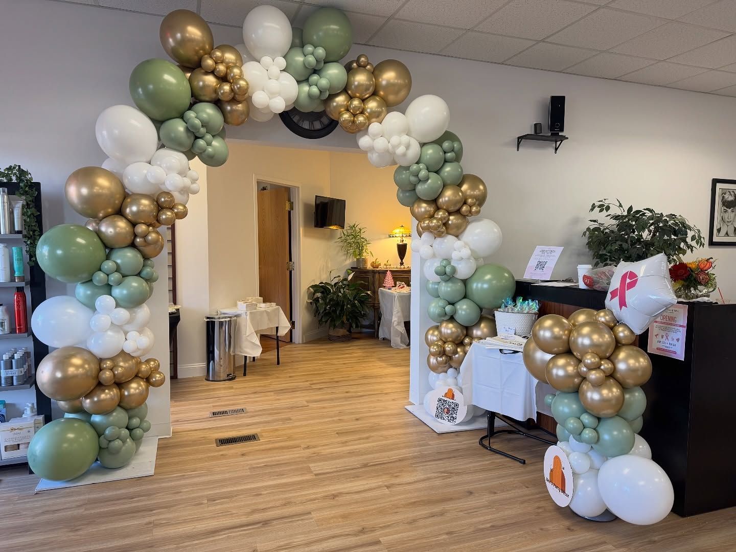 Balloon arch in gold, white, sage, and blue framing a doorway at an indoor event.