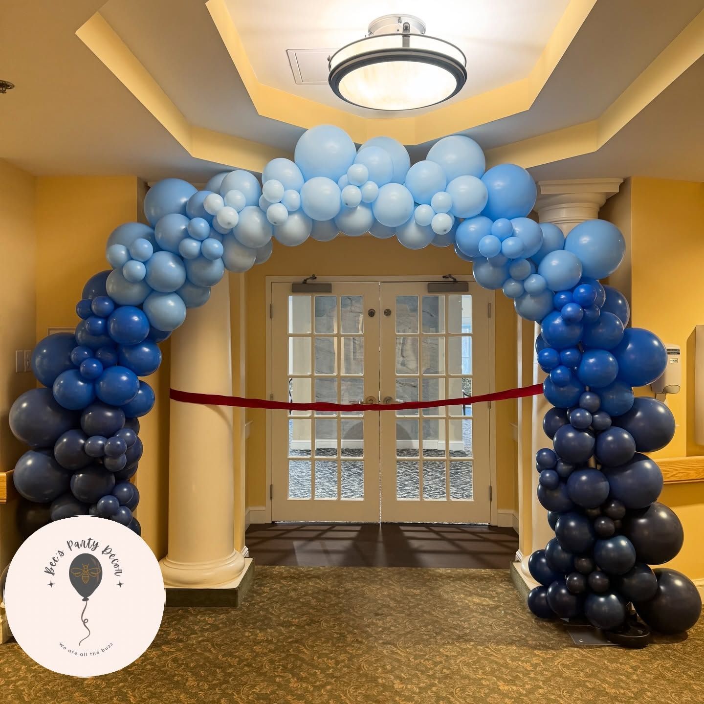 Blue balloon arch framing a doorway in a decorated hallway with a red ribbon barrier