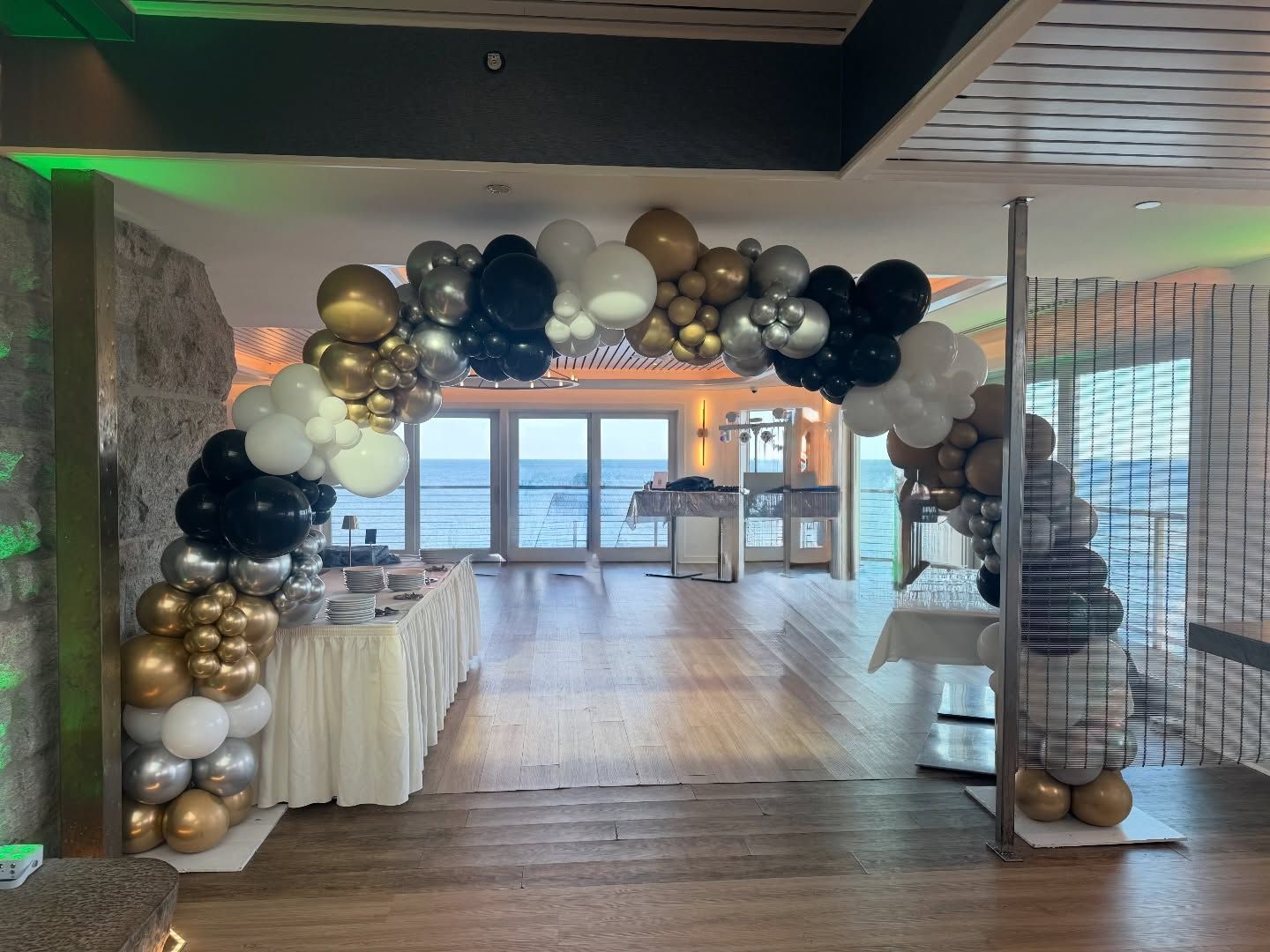 Balloon arch framing a decorated indoor event entrance with gold, black, and white balloons and table displays