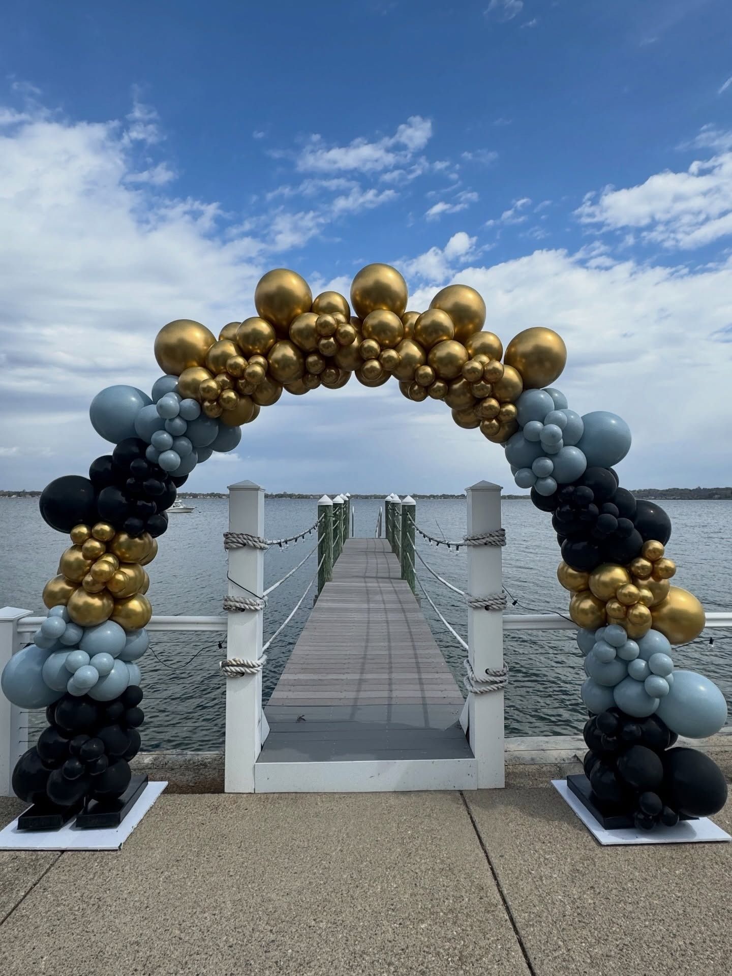 Black, gold, and silver balloon arch framing a seaside pier under a blue sky