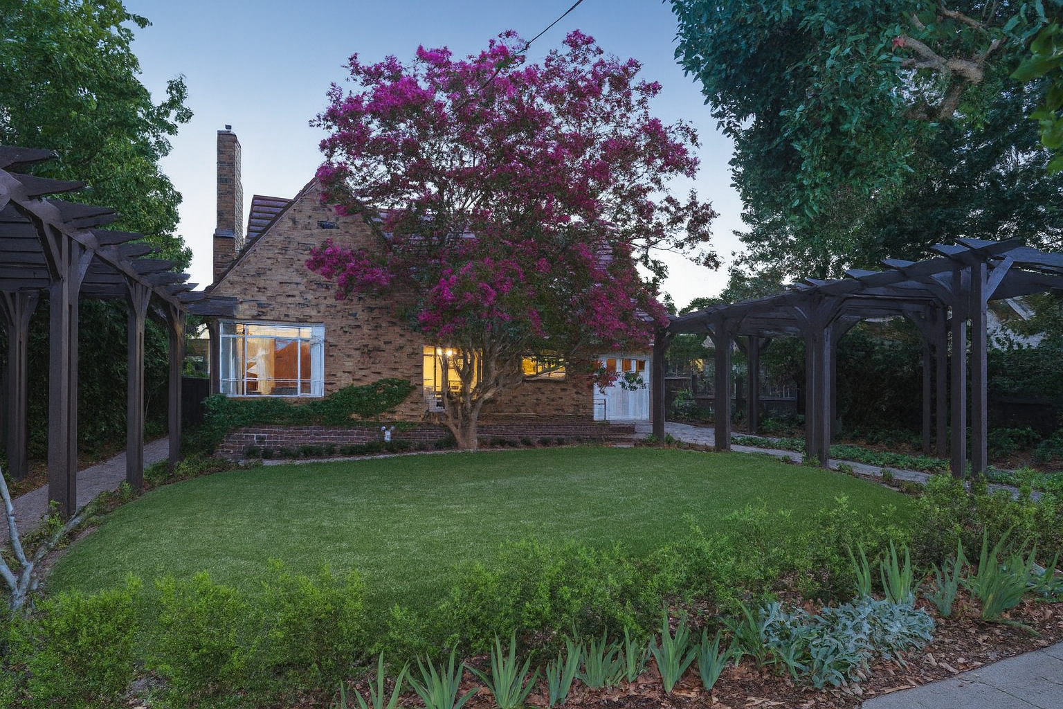 Stone cottage with a pink-flowered tree in front, flanked by garden beds and wooden trellises.