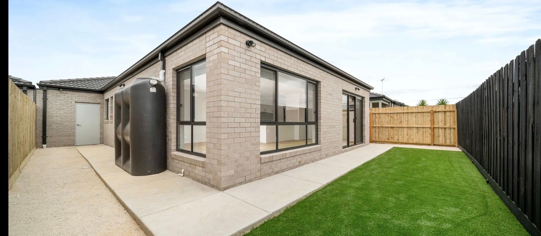 Exterior view of a modern brick house with a green lawn, black fence, and water tank.