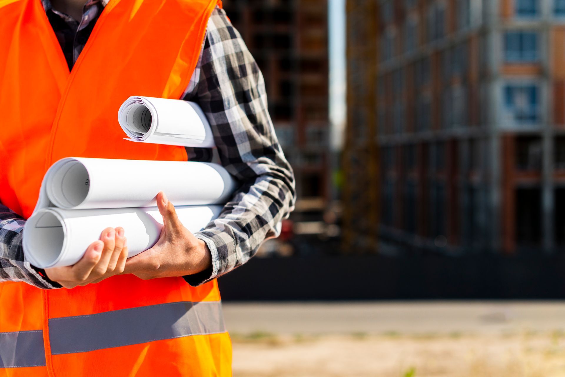 Construction worker holding rolled blueprints, wearing an orange safety vest, building in background.