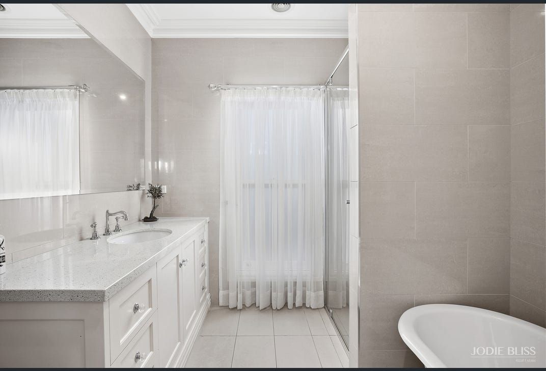 Bathroom with white vanity, tub, and window with sheer curtains. Light beige walls.