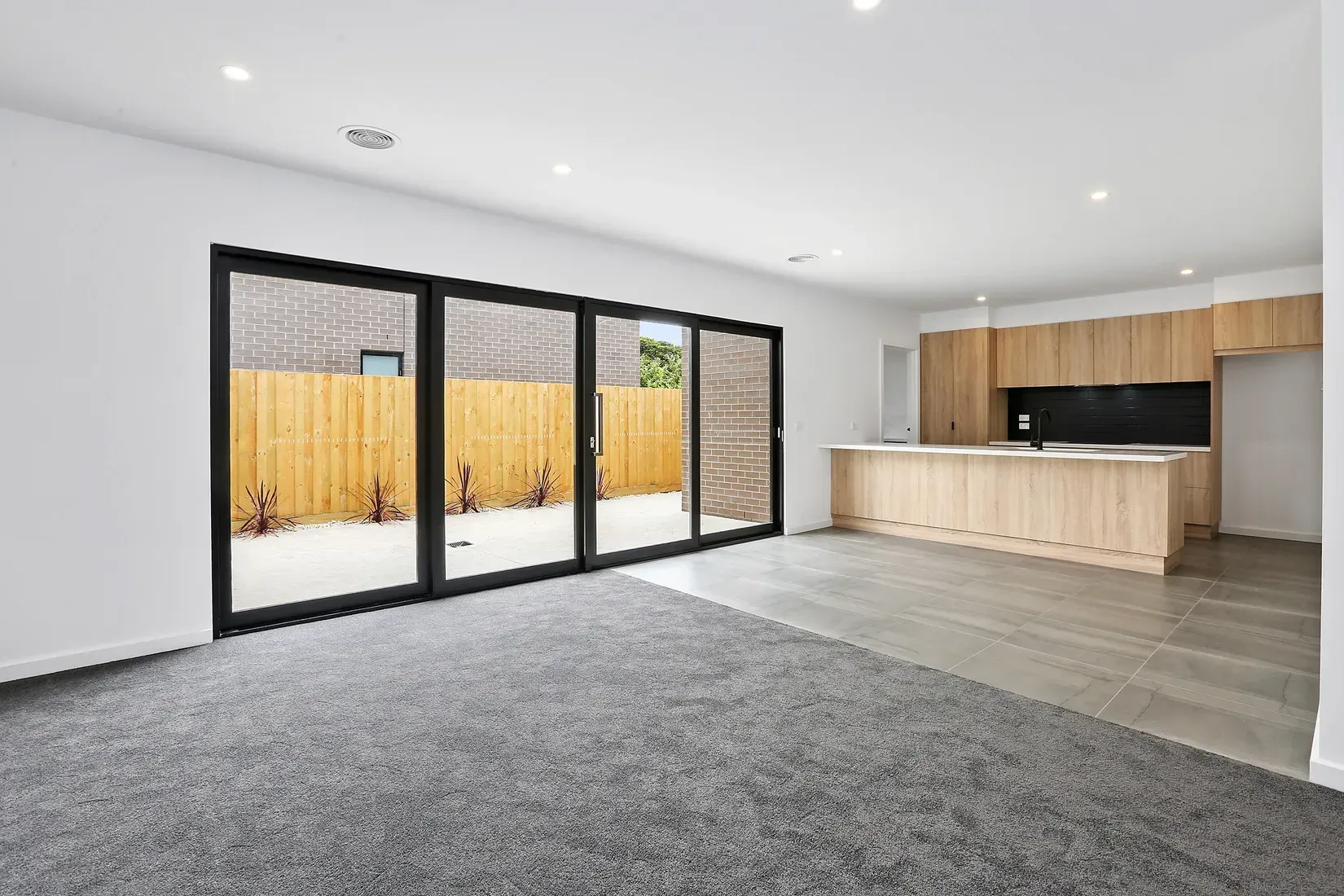 Empty living space with gray carpet, sliding glass doors, and light wood kitchen cabinets.