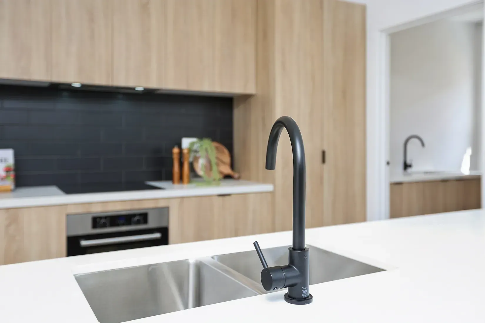 Modern kitchen with black faucet, stainless steel sink, light wood cabinets, and white countertop.