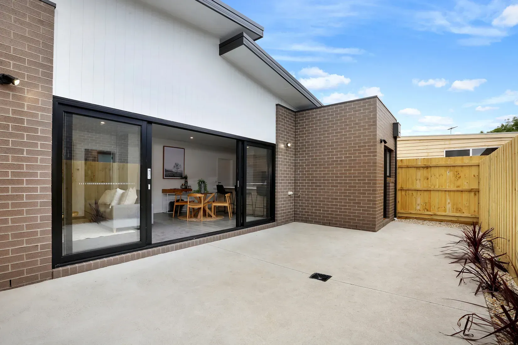 Exterior patio with sliding glass doors, brick walls, and a wooden fence under a blue sky.