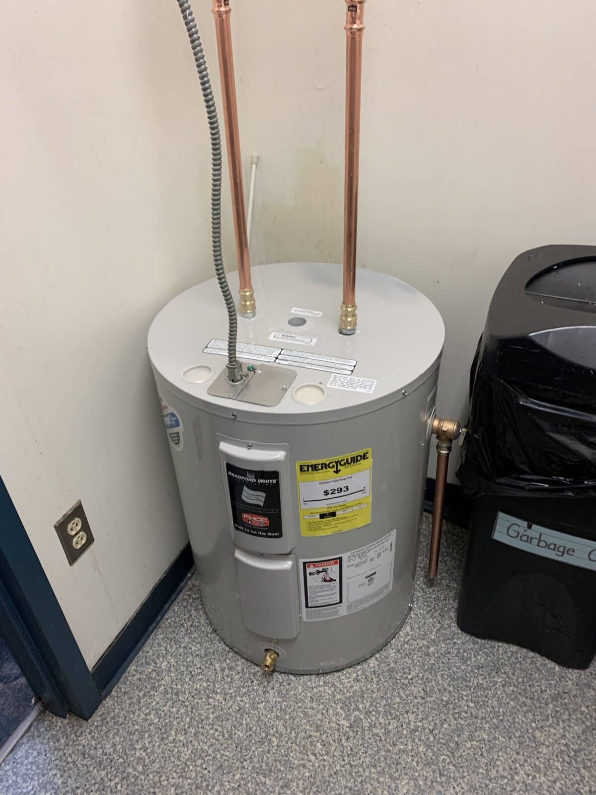 A water heater is sitting on the floor in a room next to a trash can.