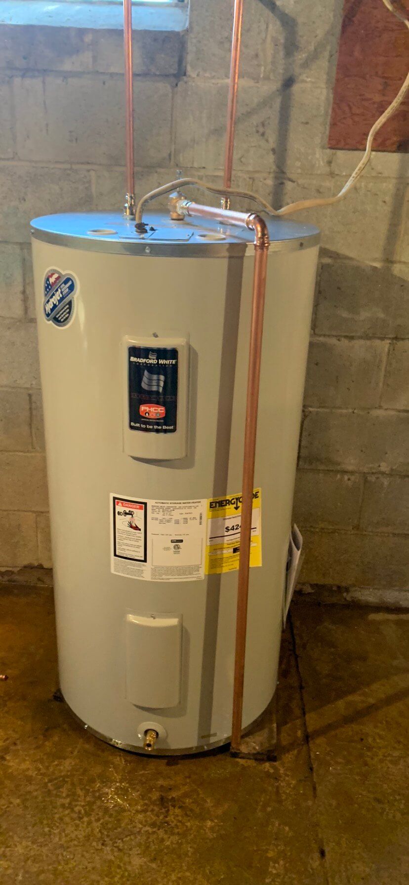 A water heater is sitting in a basement next to a brick wall.