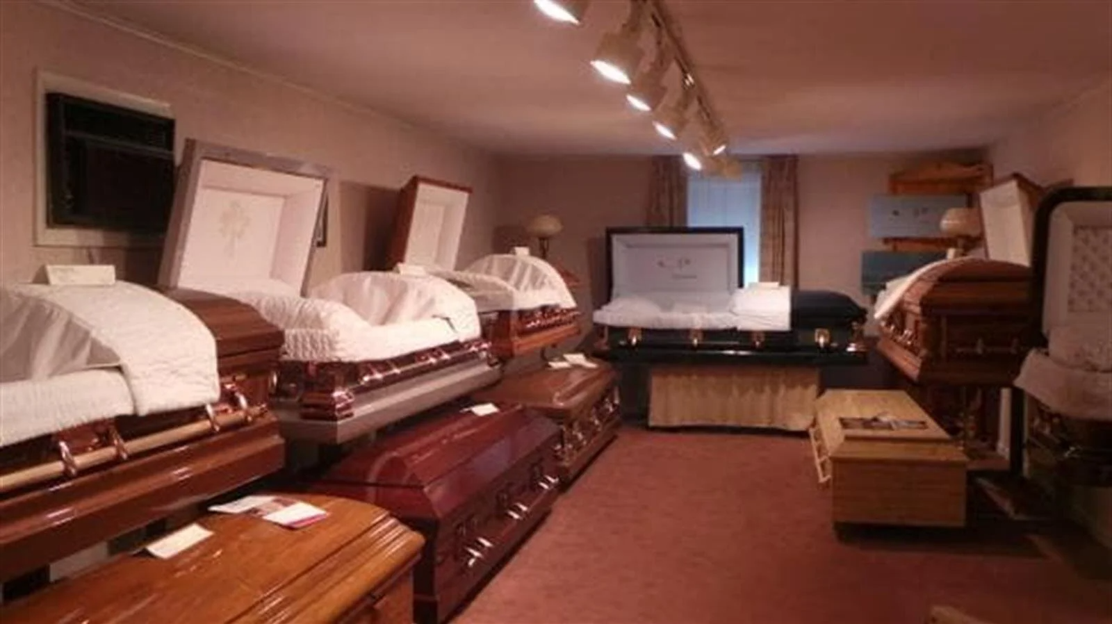 Funeral home showroom displaying various closed and open caskets; wood and metal finishes, upholstered interiors.