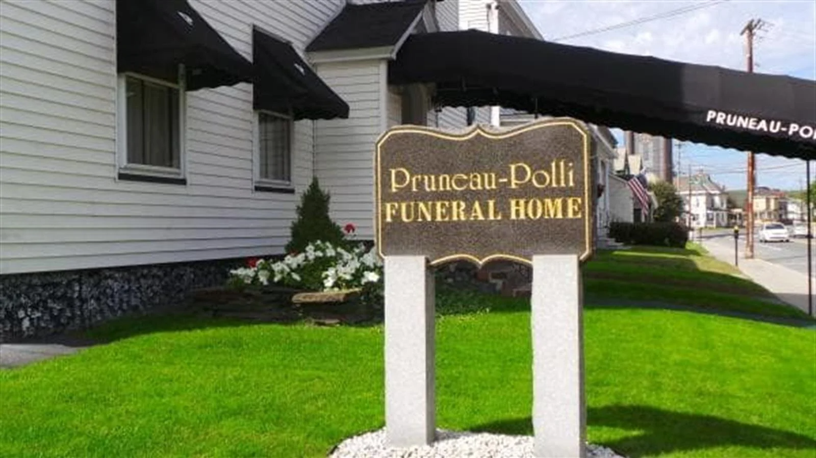 Sign for Pruneau-Polli Funeral Home, a white building with black awnings, on a grassy lawn.
