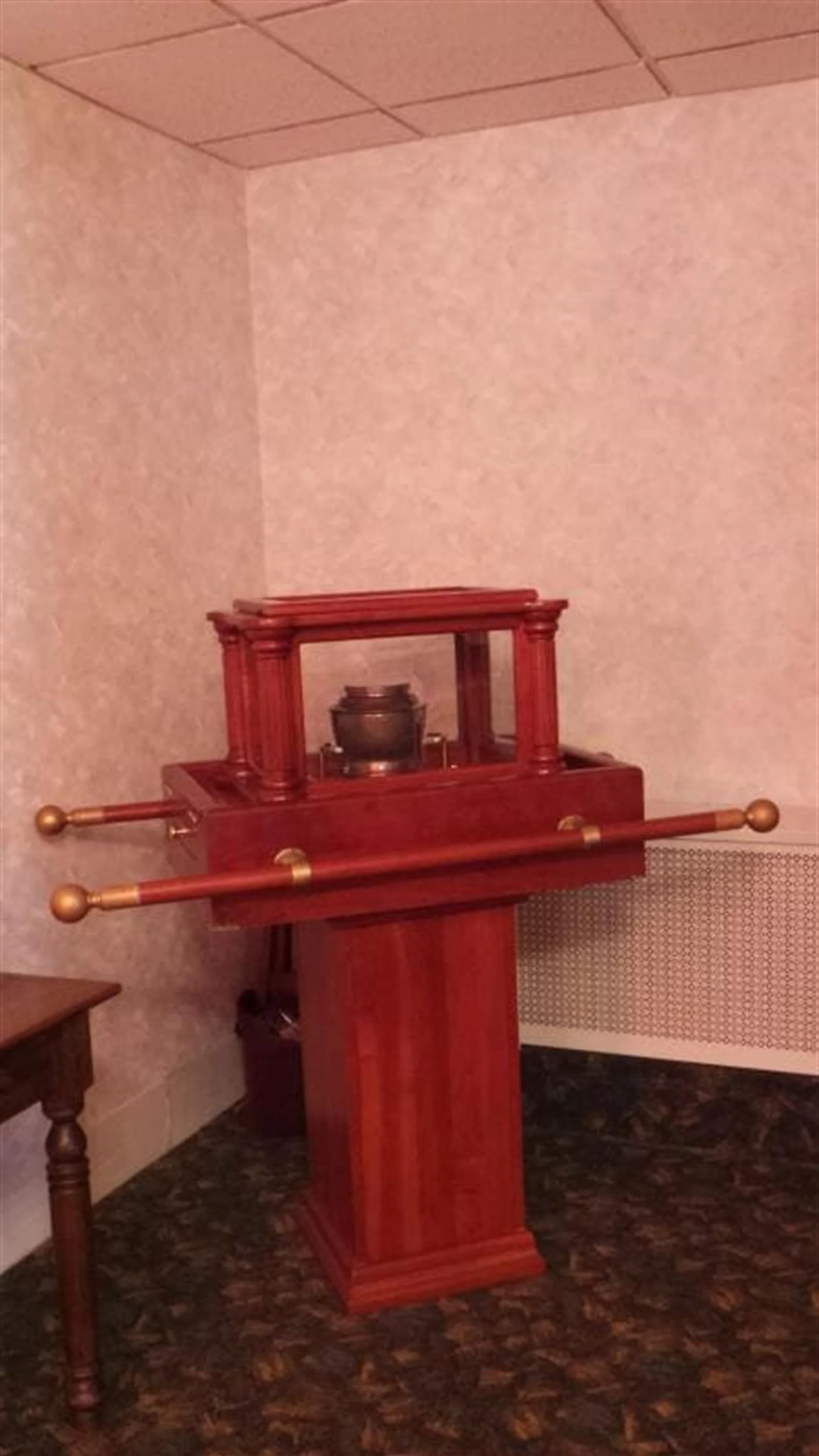 Red wooden display with a vessel on top, in a room with pale walls and a small table.