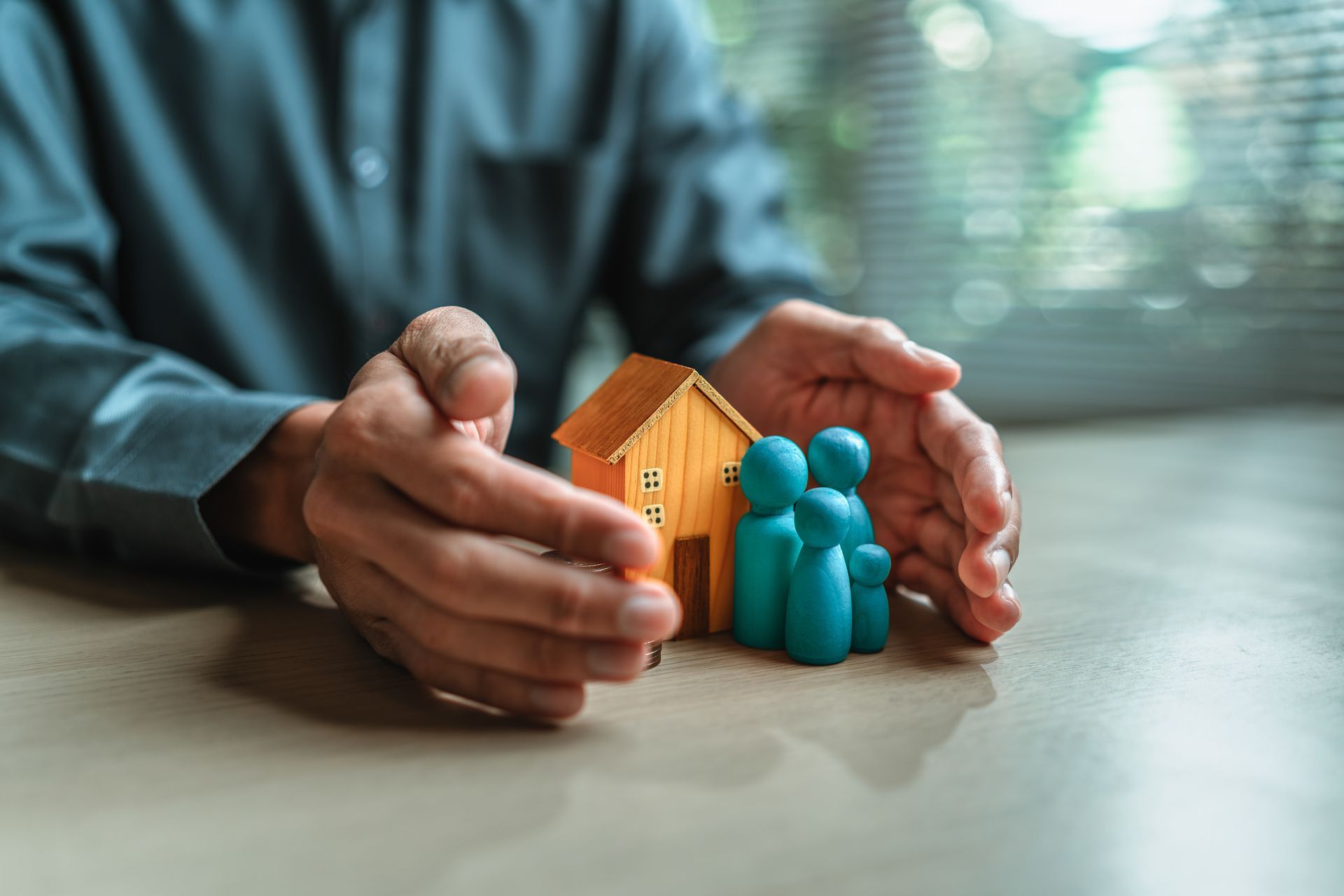 An insurance agent’s hands around a model home and wooden dolls representing a family. An insurance agent’s hands around a model home and wooden dolls representing a family.