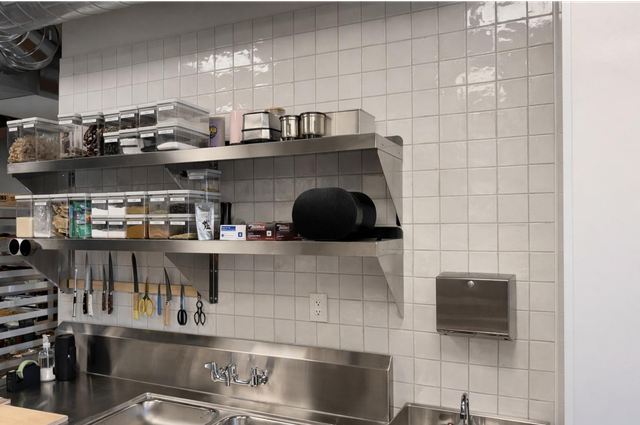 Stainless-steel kitchen with white tile wall, shelves of cookware, and a sink below.