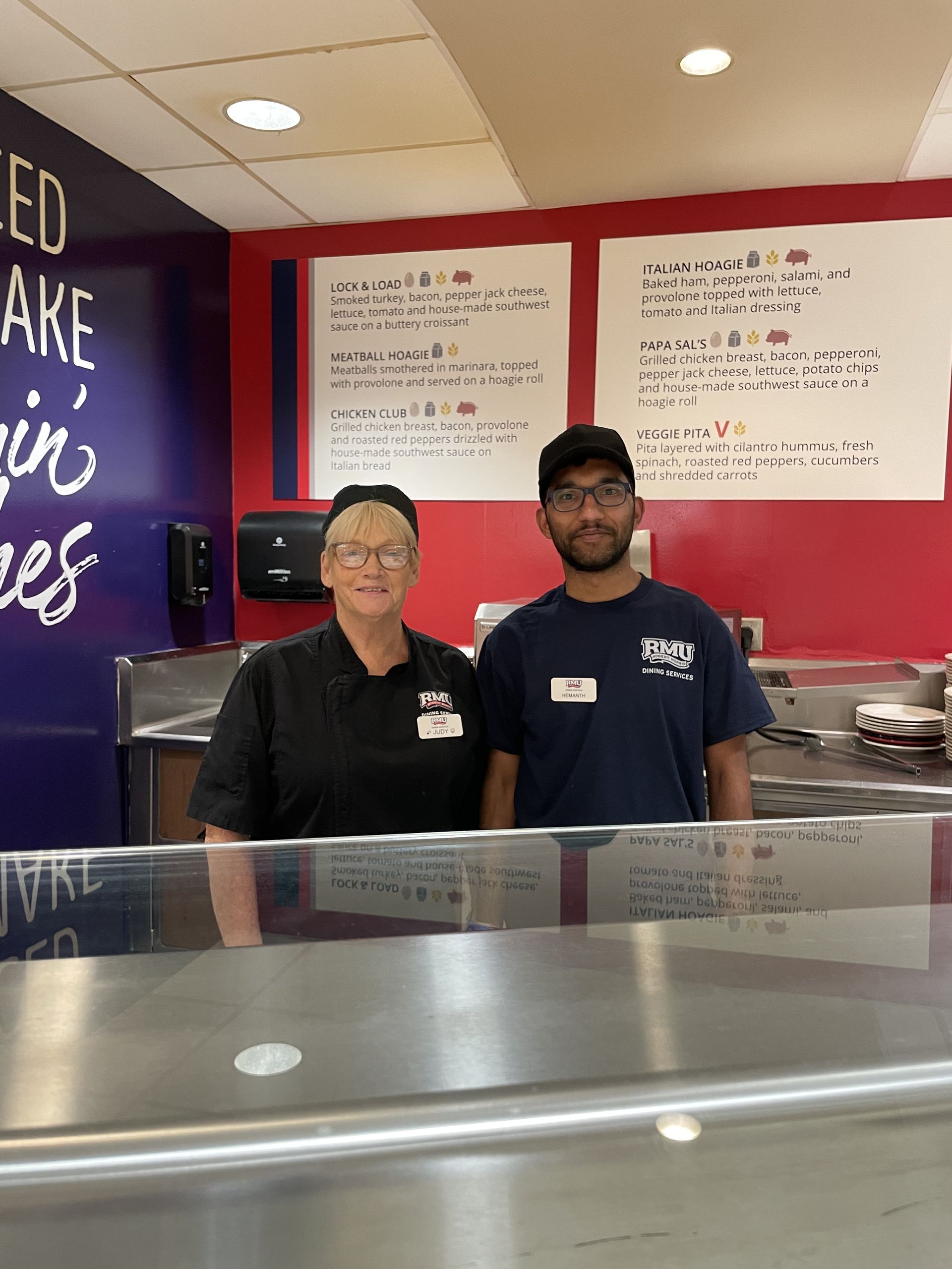 Two restaurant workers behind a counter; one woman in black, one man in a blue shirt. Red and blue wall behind them.