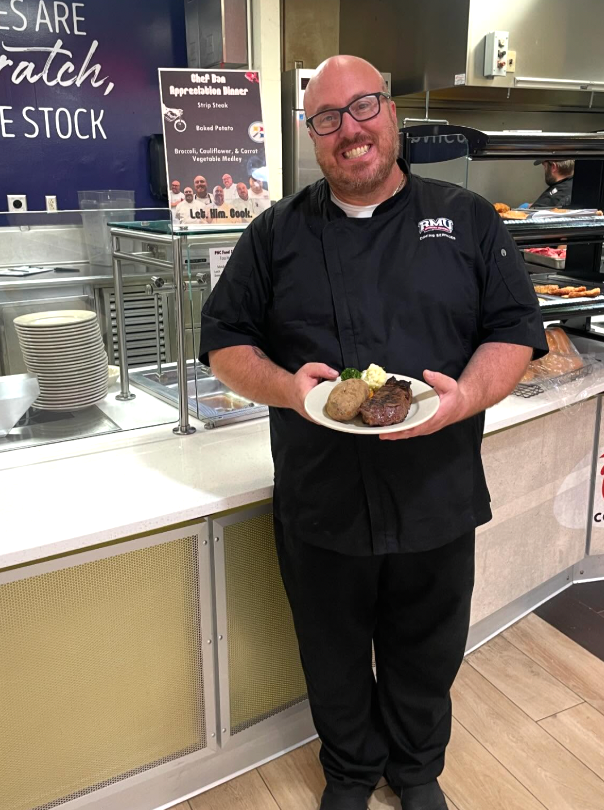 A chef in a black uniform smiles while holding a plate with steak and potatoes. He stands in a cafeteria.