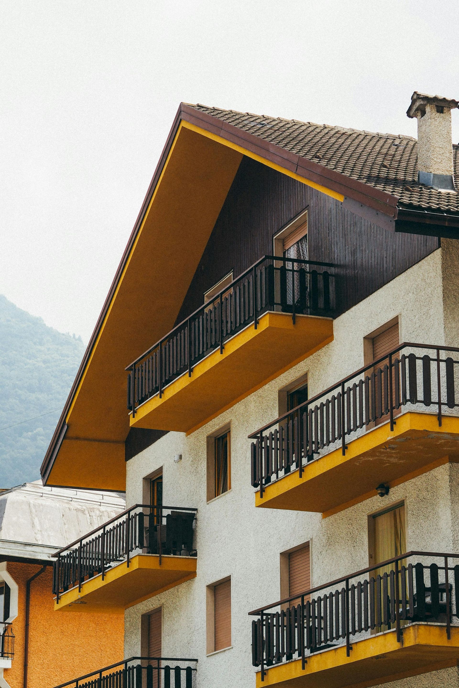 A multi-story building featuring textured white walls, yellow balconies with black railings, and a peaked wooden roof.